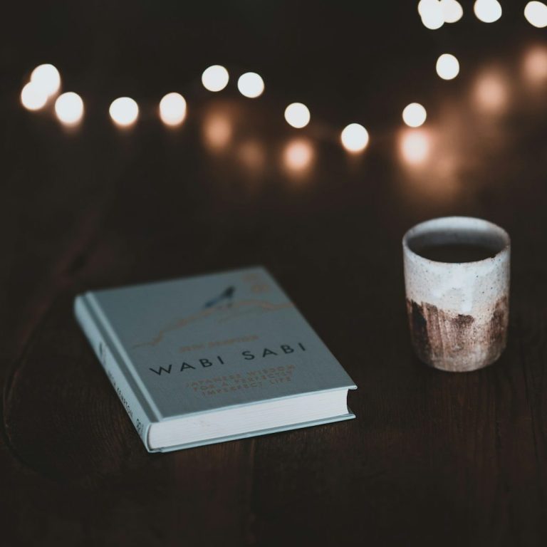 Ein Buch mit dem Titel "Wabi Sabi" und eine Tasse auf einem Holztisch, umgeben von Lichtern.