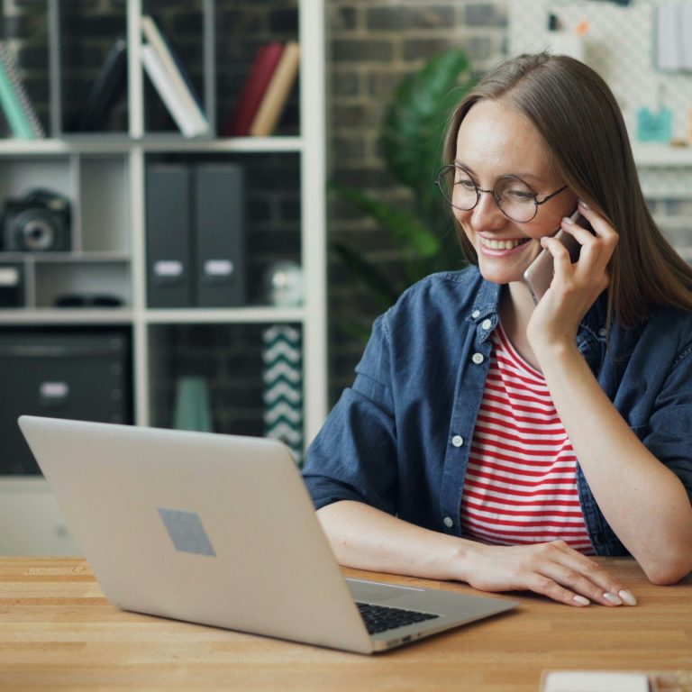 Frau mit Laptop auf einem Schreibtisch und einen Telefon in der Hand. 