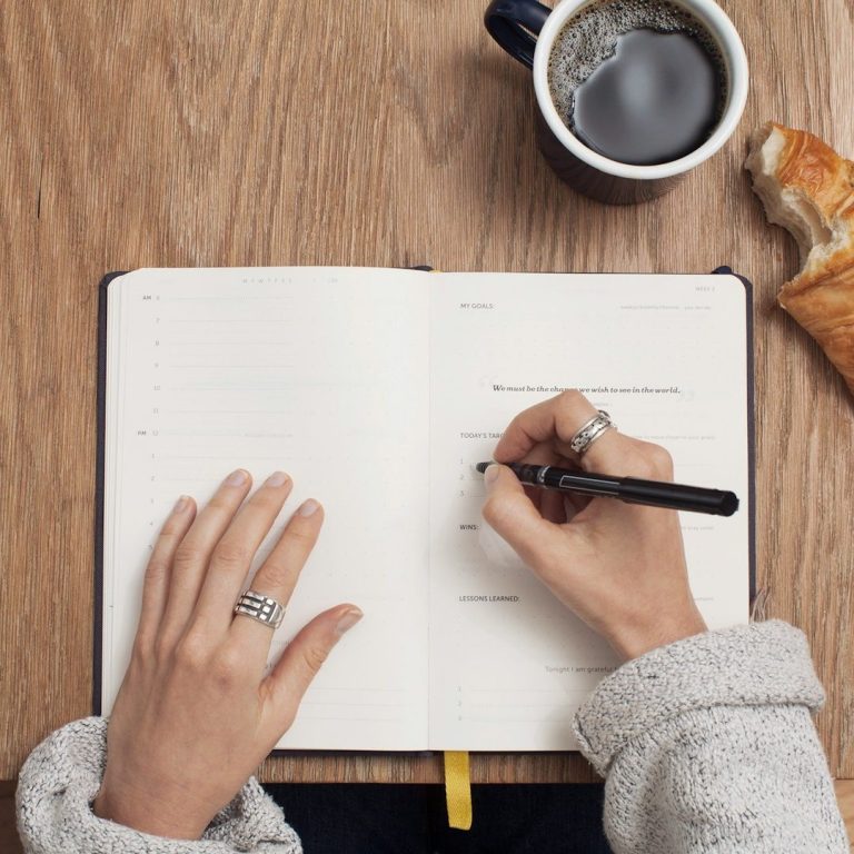 Hände schreiben in ein Notizbuch neben einer Tasse Kaffee und einem Stück Gebäck.